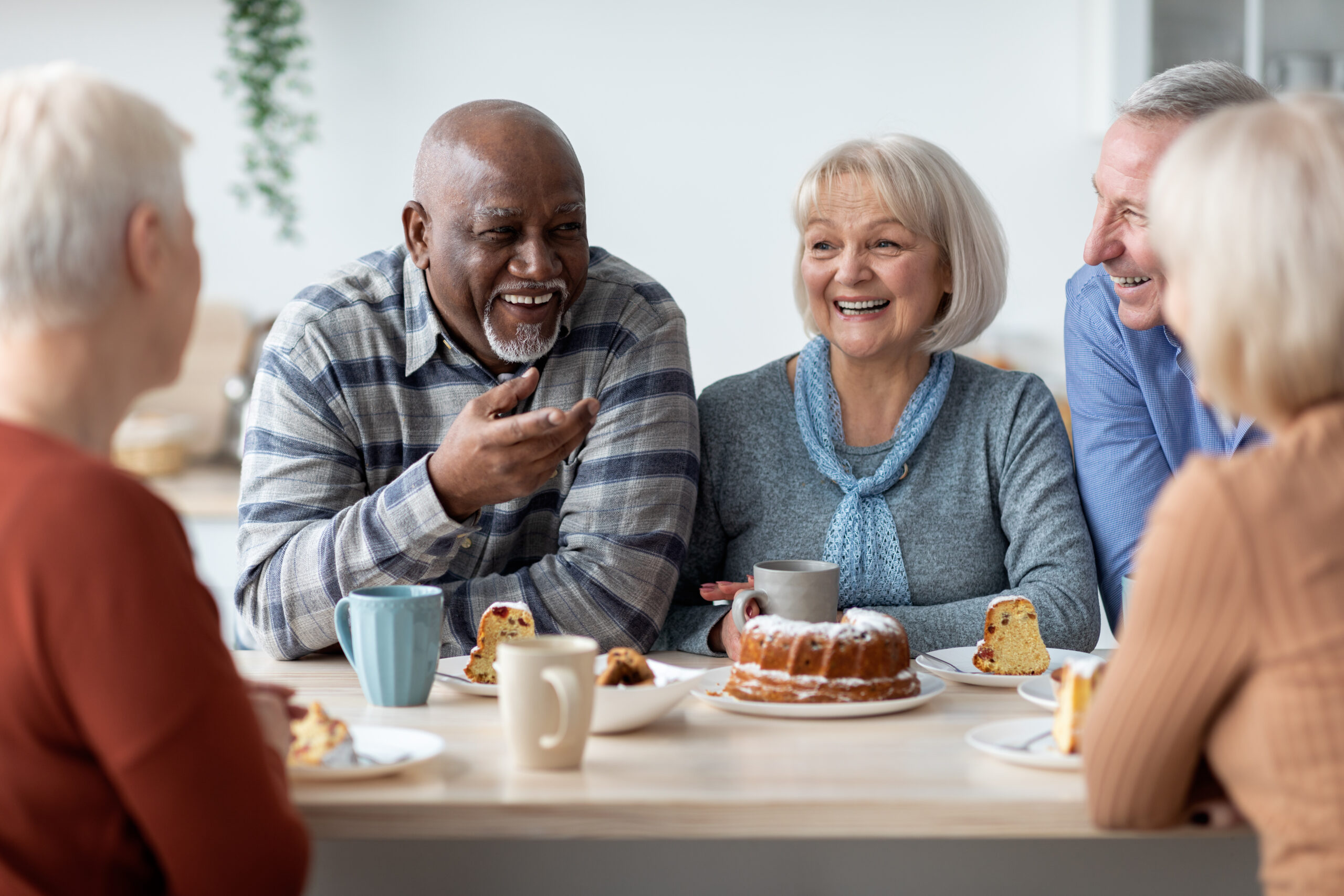 International group of positive men and women sitting around table drinking tea with cake and having conversation, smiling and laughing, having home party or enjoying time at nursing home